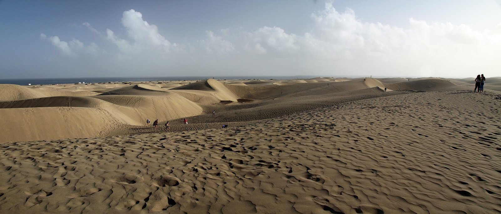 Weite Sandlandschaft der Dünen von Maspalomas mit Spaziergängern und dem Atlantik im Hintergrund.